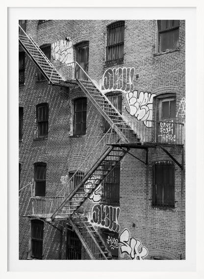 A black and white photograph of a brick apartment building with a prominent metal fire escape zigzagging across its facade. The walls are adorned with various styles of graffiti, and the windows are covered with bars, creating a gritty, urban scene. Decor