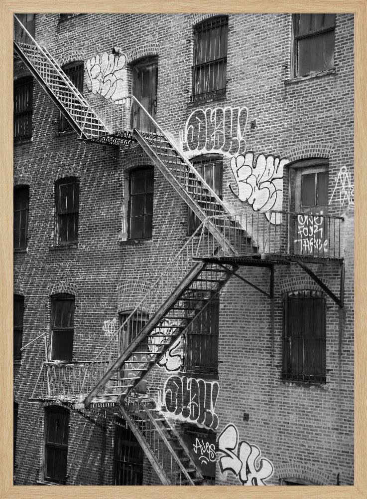 A black and white photograph of a brick apartment building with a prominent metal fire escape zigzagging across its facade. The walls are adorned with various styles of graffiti, and the windows are covered with bars, creating a gritty, urban scene. Decor
