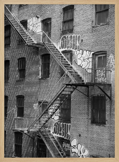 A black and white photograph of a brick apartment building with a prominent metal fire escape zigzagging across its facade. The walls are adorned with various styles of graffiti, and the windows are covered with bars, creating a gritty, urban scene. Decor