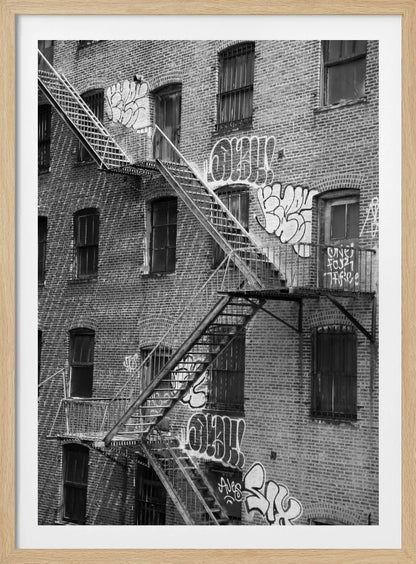 A black and white photograph of a brick apartment building with a prominent metal fire escape zigzagging across its facade. The walls are adorned with various styles of graffiti, and the windows are covered with bars, creating a gritty, urban scene. Decor