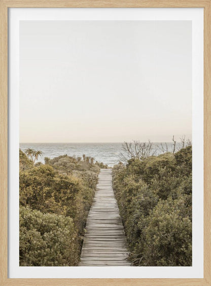 A vertical photograph of a weathered wooden boardwalk leading through dense coastal greenery towards the ocean under a pale, hazy sky, framed in black. Wall Art