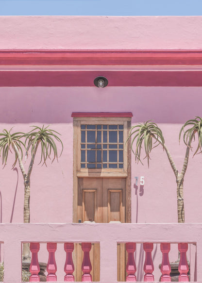 A brightly colored pink building stands under a clear blue sky, framed in black. A central wooden door with a paned window is flanked by two slender, palm-like trees. In the foreground, a pink balcony wall features fuchsia-colored balusters. Decor