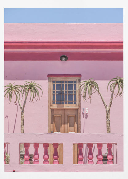 A brightly colored pink building stands under a clear blue sky, framed in black. A central wooden door with a paned window is flanked by two slender, palm-like trees. In the foreground, a pink balcony wall features fuchsia-colored balusters. Decor