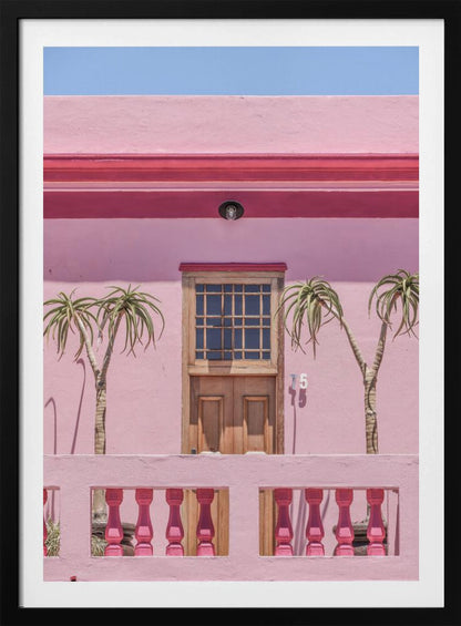 A brightly colored pink building stands under a clear blue sky, framed in black. A central wooden door with a paned window is flanked by two slender, palm-like trees. In the foreground, a pink balcony wall features fuchsia-colored balusters. Decor