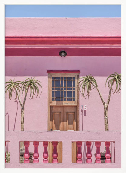 A brightly colored pink building stands under a clear blue sky, framed in black. A central wooden door with a paned window is flanked by two slender, palm-like trees. In the foreground, a pink balcony wall features fuchsia-colored balusters. Decor
