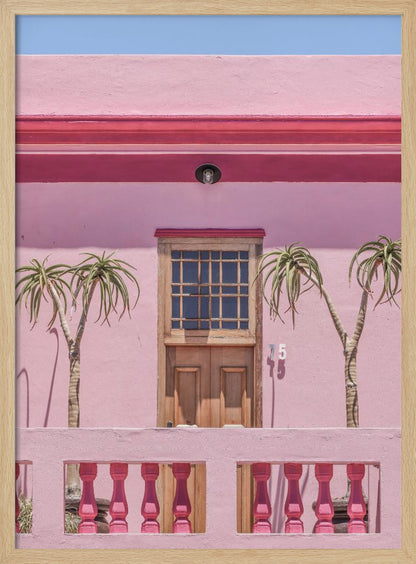 A brightly colored pink building stands under a clear blue sky, framed in black. A central wooden door with a paned window is flanked by two slender, palm-like trees. In the foreground, a pink balcony wall features fuchsia-colored balusters. Decor