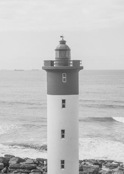 A framed, black and white photograph of a tall, cylindrical lighthouse standing on a rocky shore. The lighthouse is dark grey on its upper section and white on the lower, with the ocean and a grey sky in the background. Print