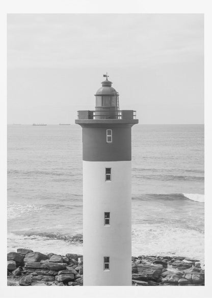 A framed, black and white photograph of a tall, cylindrical lighthouse standing on a rocky shore. The lighthouse is dark grey on its upper section and white on the lower, with the ocean and a grey sky in the background. Print