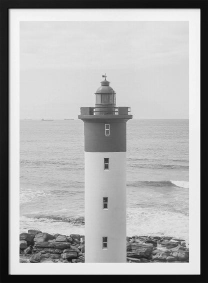 A framed, black and white photograph of a tall, cylindrical lighthouse standing on a rocky shore. The lighthouse is dark grey on its upper section and white on the lower, with the ocean and a grey sky in the background. Print