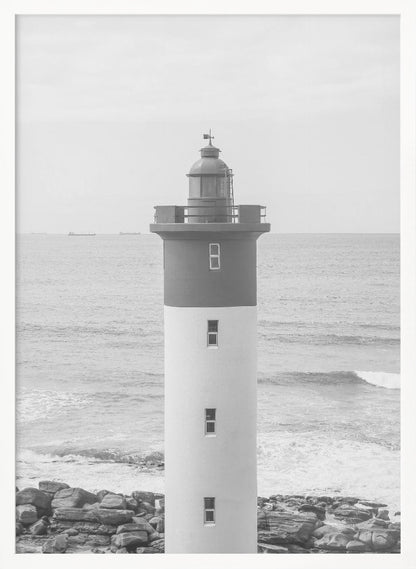 A framed, black and white photograph of a tall, cylindrical lighthouse standing on a rocky shore. The lighthouse is dark grey on its upper section and white on the lower, with the ocean and a grey sky in the background. Print