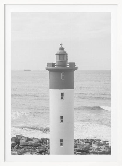 A framed, black and white photograph of a tall, cylindrical lighthouse standing on a rocky shore. The lighthouse is dark grey on its upper section and white on the lower, with the ocean and a grey sky in the background. Print