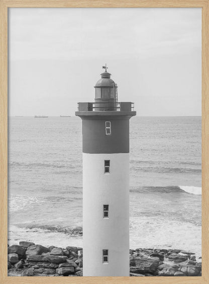 A framed, black and white photograph of a tall, cylindrical lighthouse standing on a rocky shore. The lighthouse is dark grey on its upper section and white on the lower, with the ocean and a grey sky in the background. Print
