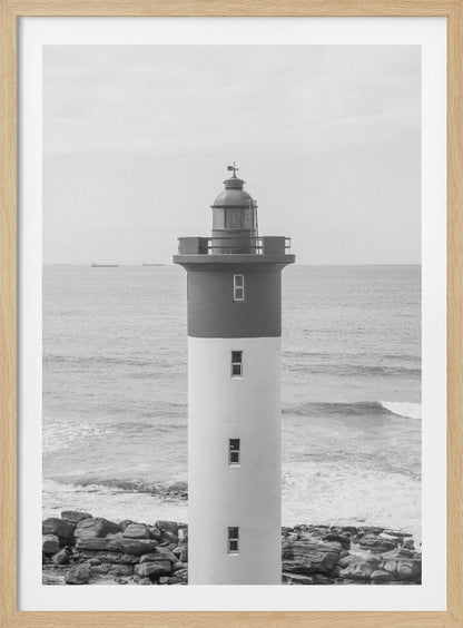 A framed, black and white photograph of a tall, cylindrical lighthouse standing on a rocky shore. The lighthouse is dark grey on its upper section and white on the lower, with the ocean and a grey sky in the background. Print