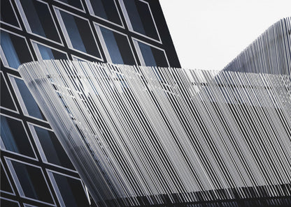 An abstract architectural photograph, framed in silver, showing a dynamic contrast between two modern building facades. In the background, a dark building features a grid of diamond-shaped windows with white frames, while a structure in the foreground creates a wave of fine, parallel white vertical lines against the sky and the other building. Print