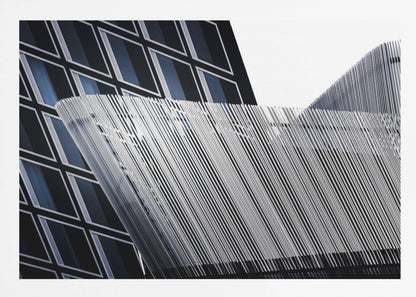 An abstract architectural photograph, framed in silver, showing a dynamic contrast between two modern building facades. In the background, a dark building features a grid of diamond-shaped windows with white frames, while a structure in the foreground creates a wave of fine, parallel white vertical lines against the sky and the other building. Print