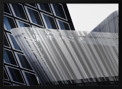 An abstract architectural photograph, framed in silver, showing a dynamic contrast between two modern building facades. In the background, a dark building features a grid of diamond-shaped windows with white frames, while a structure in the foreground creates a wave of fine, parallel white vertical lines against the sky and the other building. Print