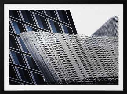 An abstract architectural photograph, framed in silver, showing a dynamic contrast between two modern building facades. In the background, a dark building features a grid of diamond-shaped windows with white frames, while a structure in the foreground creates a wave of fine, parallel white vertical lines against the sky and the other building. Print