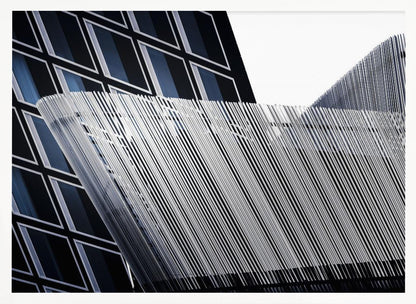 An abstract architectural photograph, framed in silver, showing a dynamic contrast between two modern building facades. In the background, a dark building features a grid of diamond-shaped windows with white frames, while a structure in the foreground creates a wave of fine, parallel white vertical lines against the sky and the other building. Print
