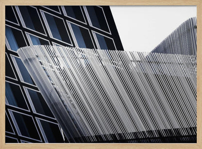 An abstract architectural photograph, framed in silver, showing a dynamic contrast between two modern building facades. In the background, a dark building features a grid of diamond-shaped windows with white frames, while a structure in the foreground creates a wave of fine, parallel white vertical lines against the sky and the other building. Print