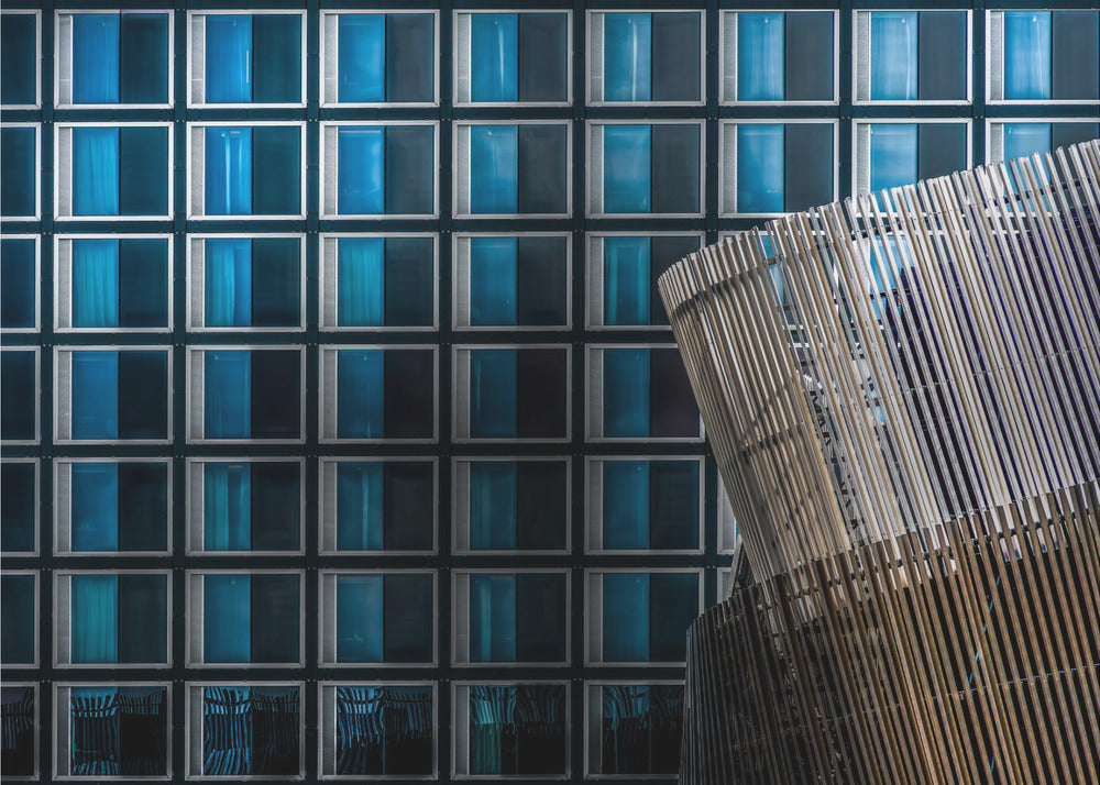 An architectural abstract photograph featuring a modern building facade with a grid of blue and black square windows, contrasted by a curved, slatted structure in the foreground, all enclosed in a silver frame. Wall Art