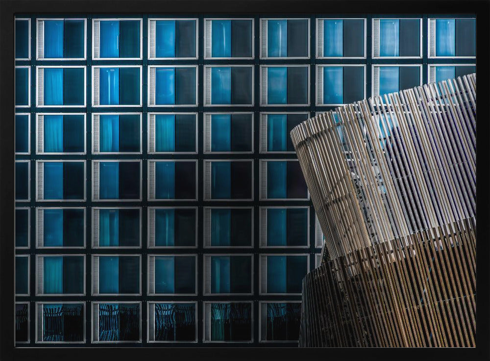 An architectural abstract photograph featuring a modern building facade with a grid of blue and black square windows, contrasted by a curved, slatted structure in the foreground, all enclosed in a silver frame. Wall Art