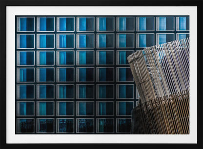 An architectural abstract photograph featuring a modern building facade with a grid of blue and black square windows, contrasted by a curved, slatted structure in the foreground, all enclosed in a silver frame. Wall Art