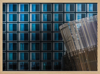 An architectural abstract photograph featuring a modern building facade with a grid of blue and black square windows, contrasted by a curved, slatted structure in the foreground, all enclosed in a silver frame. Wall Art