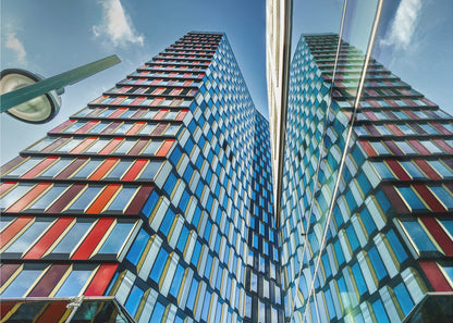 A low-angle shot of a modern skyscraper with a colorful facade of blue, red, and orange geometric panels, reflected in an adjacent glass building under a bright blue sky, all enclosed in a silver frame. Poster
