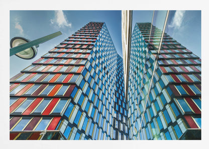 A low-angle shot of a modern skyscraper with a colorful facade of blue, red, and orange geometric panels, reflected in an adjacent glass building under a bright blue sky, all enclosed in a silver frame. Poster
