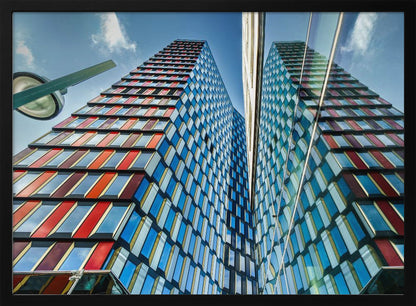 A low-angle shot of a modern skyscraper with a colorful facade of blue, red, and orange geometric panels, reflected in an adjacent glass building under a bright blue sky, all enclosed in a silver frame. Poster