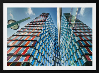 A low-angle shot of a modern skyscraper with a colorful facade of blue, red, and orange geometric panels, reflected in an adjacent glass building under a bright blue sky, all enclosed in a silver frame. Poster
