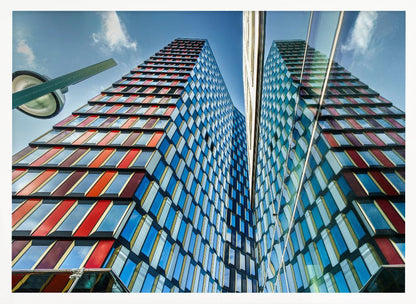 A low-angle shot of a modern skyscraper with a colorful facade of blue, red, and orange geometric panels, reflected in an adjacent glass building under a bright blue sky, all enclosed in a silver frame. Poster