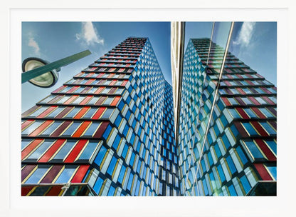 A low-angle shot of a modern skyscraper with a colorful facade of blue, red, and orange geometric panels, reflected in an adjacent glass building under a bright blue sky, all enclosed in a silver frame. Poster