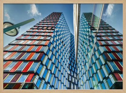 A low-angle shot of a modern skyscraper with a colorful facade of blue, red, and orange geometric panels, reflected in an adjacent glass building under a bright blue sky, all enclosed in a silver frame. Poster