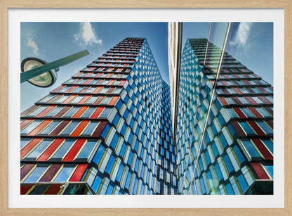 A low-angle shot of a modern skyscraper with a colorful facade of blue, red, and orange geometric panels, reflected in an adjacent glass building under a bright blue sky, all enclosed in a silver frame. Poster