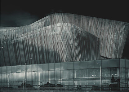 A dramatic black and white photograph of a modern building with a unique, wave-like facade made of vertical fins. The lower level is made of glass, reflecting the sky and streetlights. The entire image is presented in a silver frame. Artwork