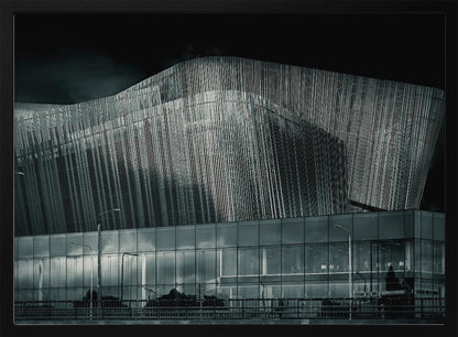 A dramatic black and white photograph of a modern building with a unique, wave-like facade made of vertical fins. The lower level is made of glass, reflecting the sky and streetlights. The entire image is presented in a silver frame. Artwork