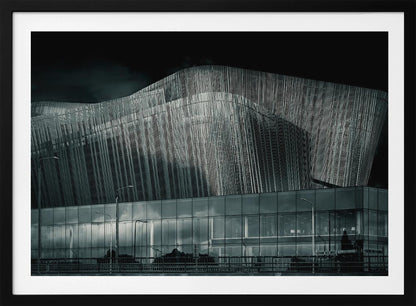 A dramatic black and white photograph of a modern building with a unique, wave-like facade made of vertical fins. The lower level is made of glass, reflecting the sky and streetlights. The entire image is presented in a silver frame. Artwork