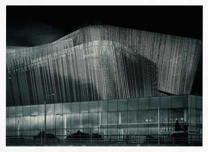 A dramatic black and white photograph of a modern building with a unique, wave-like facade made of vertical fins. The lower level is made of glass, reflecting the sky and streetlights. The entire image is presented in a silver frame. Artwork