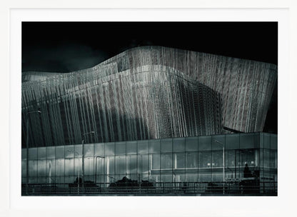 A dramatic black and white photograph of a modern building with a unique, wave-like facade made of vertical fins. The lower level is made of glass, reflecting the sky and streetlights. The entire image is presented in a silver frame. Artwork