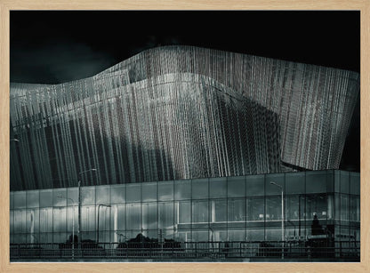 A dramatic black and white photograph of a modern building with a unique, wave-like facade made of vertical fins. The lower level is made of glass, reflecting the sky and streetlights. The entire image is presented in a silver frame. Artwork