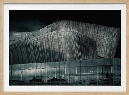 A dramatic black and white photograph of a modern building with a unique, wave-like facade made of vertical fins. The lower level is made of glass, reflecting the sky and streetlights. The entire image is presented in a silver frame. Artwork