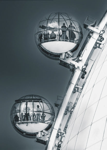 A dramatic low-angle, black and white photograph of two spherical glass pods of the London Eye Ferris wheel. Silhouettes of people are visible inside the pods against a dark, solid sky, with the massive white structure of the wheel on the right. Print