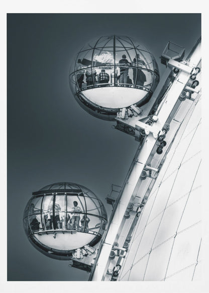 A dramatic low-angle, black and white photograph of two spherical glass pods of the London Eye Ferris wheel. Silhouettes of people are visible inside the pods against a dark, solid sky, with the massive white structure of the wheel on the right. Print
