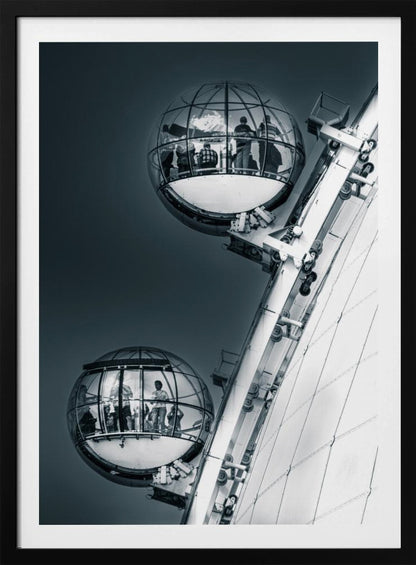 A dramatic low-angle, black and white photograph of two spherical glass pods of the London Eye Ferris wheel. Silhouettes of people are visible inside the pods against a dark, solid sky, with the massive white structure of the wheel on the right. Print