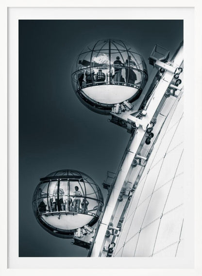 A dramatic low-angle, black and white photograph of two spherical glass pods of the London Eye Ferris wheel. Silhouettes of people are visible inside the pods against a dark, solid sky, with the massive white structure of the wheel on the right. Print