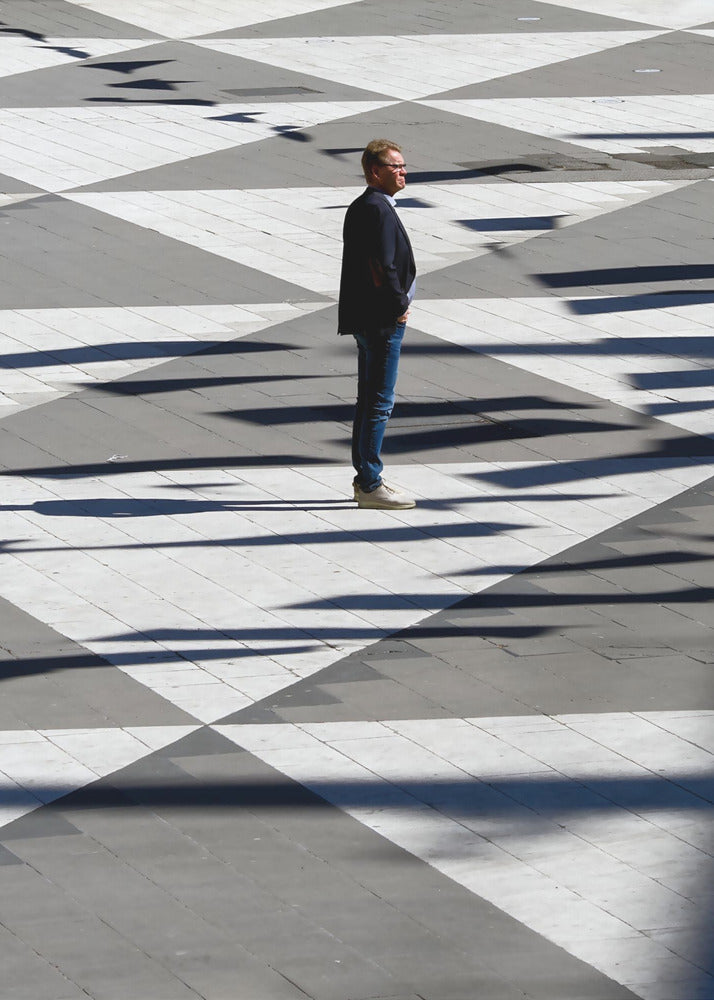 A man in a dark jacket and blue jeans stands alone in profile on a geometrically patterned plaza. The ground is a striking pattern of grey and white triangles, crossed by long, dark shadows cast by the bright sun, creating a high-contrast, graphic composition. Wall Art