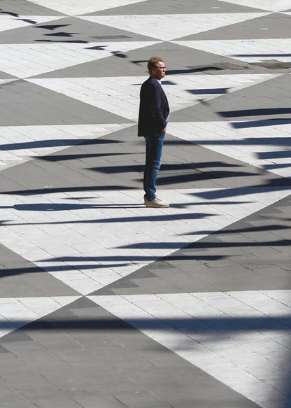 A man in a dark jacket and blue jeans stands alone in profile on a geometrically patterned plaza. The ground is a striking pattern of grey and white triangles, crossed by long, dark shadows cast by the bright sun, creating a high-contrast, graphic composition. Wall Art