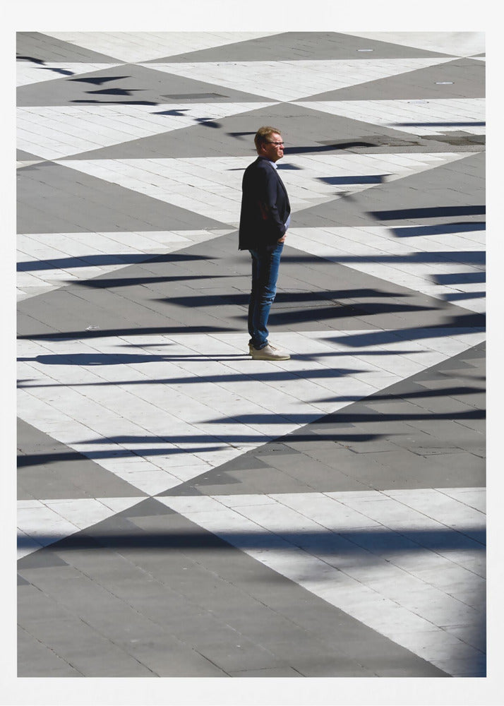 A man in a dark jacket and blue jeans stands alone in profile on a geometrically patterned plaza. The ground is a striking pattern of grey and white triangles, crossed by long, dark shadows cast by the bright sun, creating a high-contrast, graphic composition. Wall Art