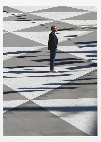 A man in a dark jacket and blue jeans stands alone in profile on a geometrically patterned plaza. The ground is a striking pattern of grey and white triangles, crossed by long, dark shadows cast by the bright sun, creating a high-contrast, graphic composition. Wall Art